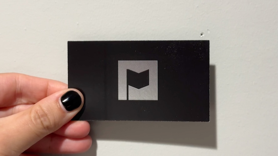 A picture of a hand holding a business card showing a stylized design of a black flag on a silver square, with a black background.