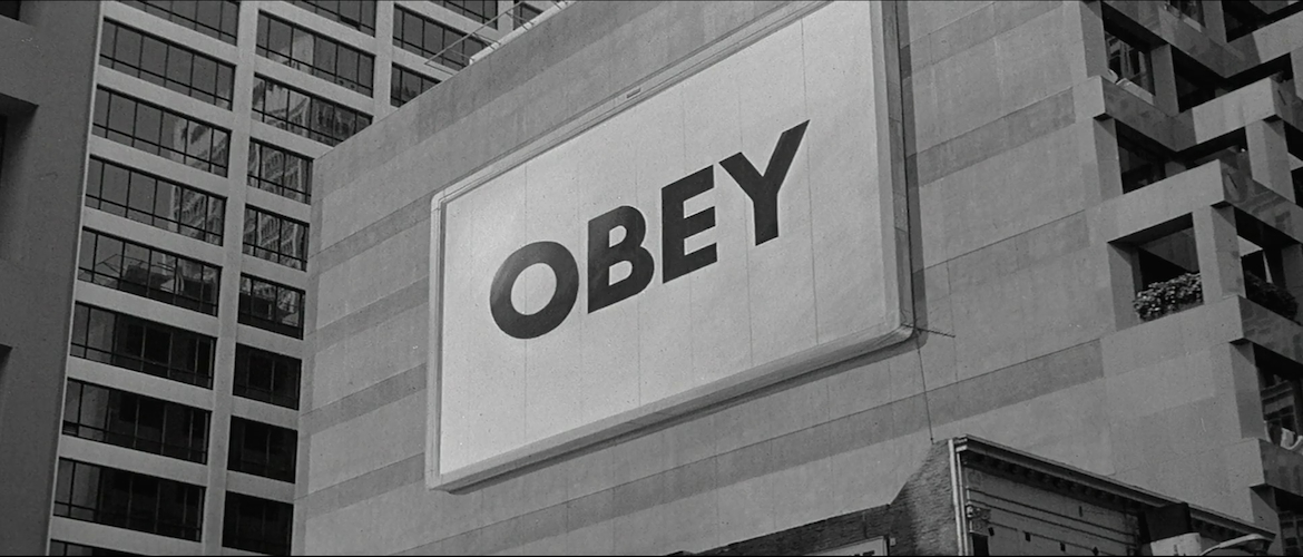 A still from the 1988 action horror film "They Live". The picture is in black and white, showing an advertising billboard on the side of an office building, showing the word OBEY.